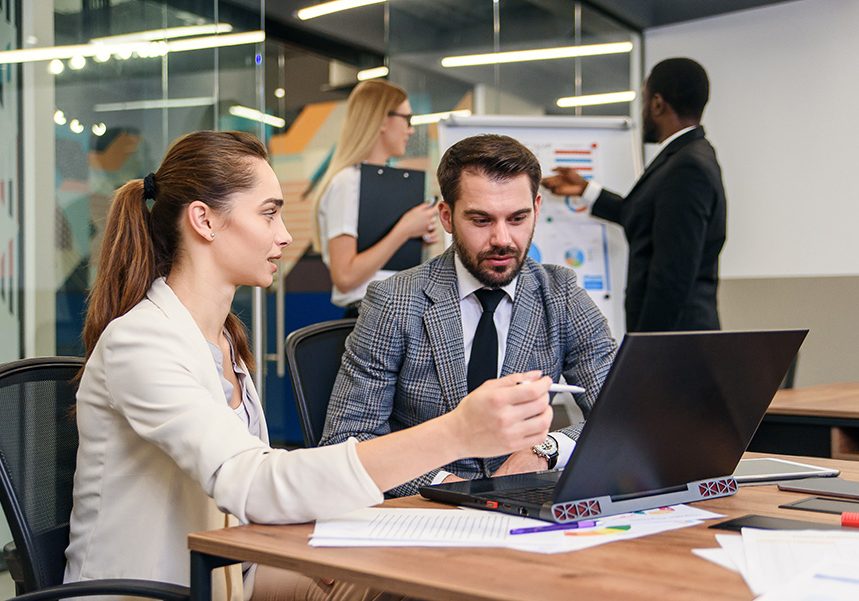 Joined work of two confident business people in formal suit working together with laptop at office with their coworkers at background. Joined work of two confident business people in formal suit working together with laptop at modern office with their coworkers at background.