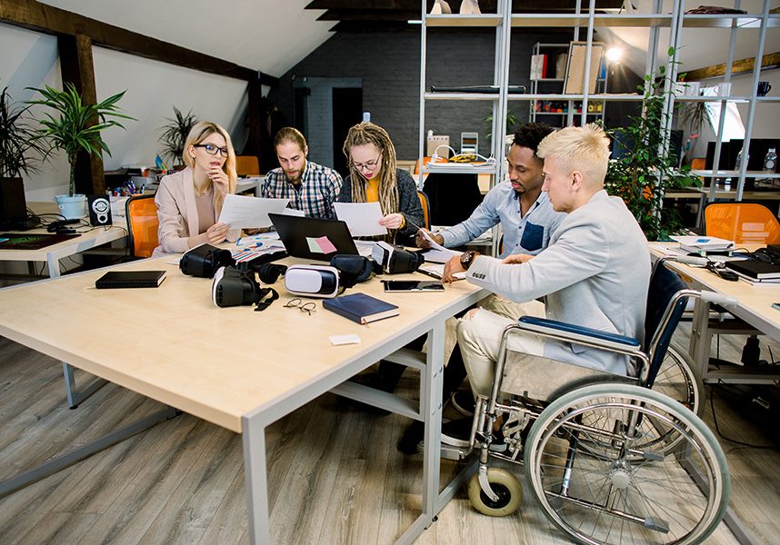 Group of five young creative multiethnic business people working together in modern office with male colleague in a wheelchair for inclusion. Successful disabled business people Group of five young creative multiethnic business people working together in modern office with male colleague in a wheelchair for inclusion. Successful disabled business people.