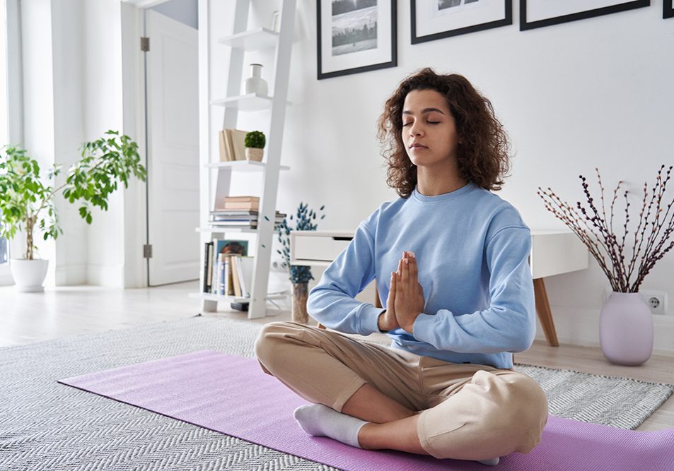 Healthy serene young woman meditating at home with eyes closed doing pilates breathing exercises, relaxing body and mind sitting on floor in living room. Mental health and meditation for no stress.