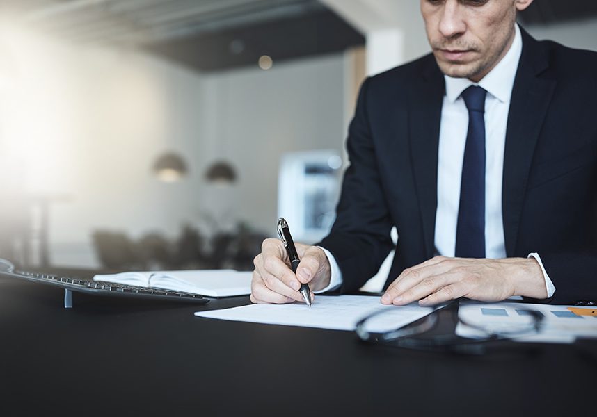 Businessman sitting at his office desk going over paperwork Focused businessman signing a document while going over paperwork at his desk in a bright modern office