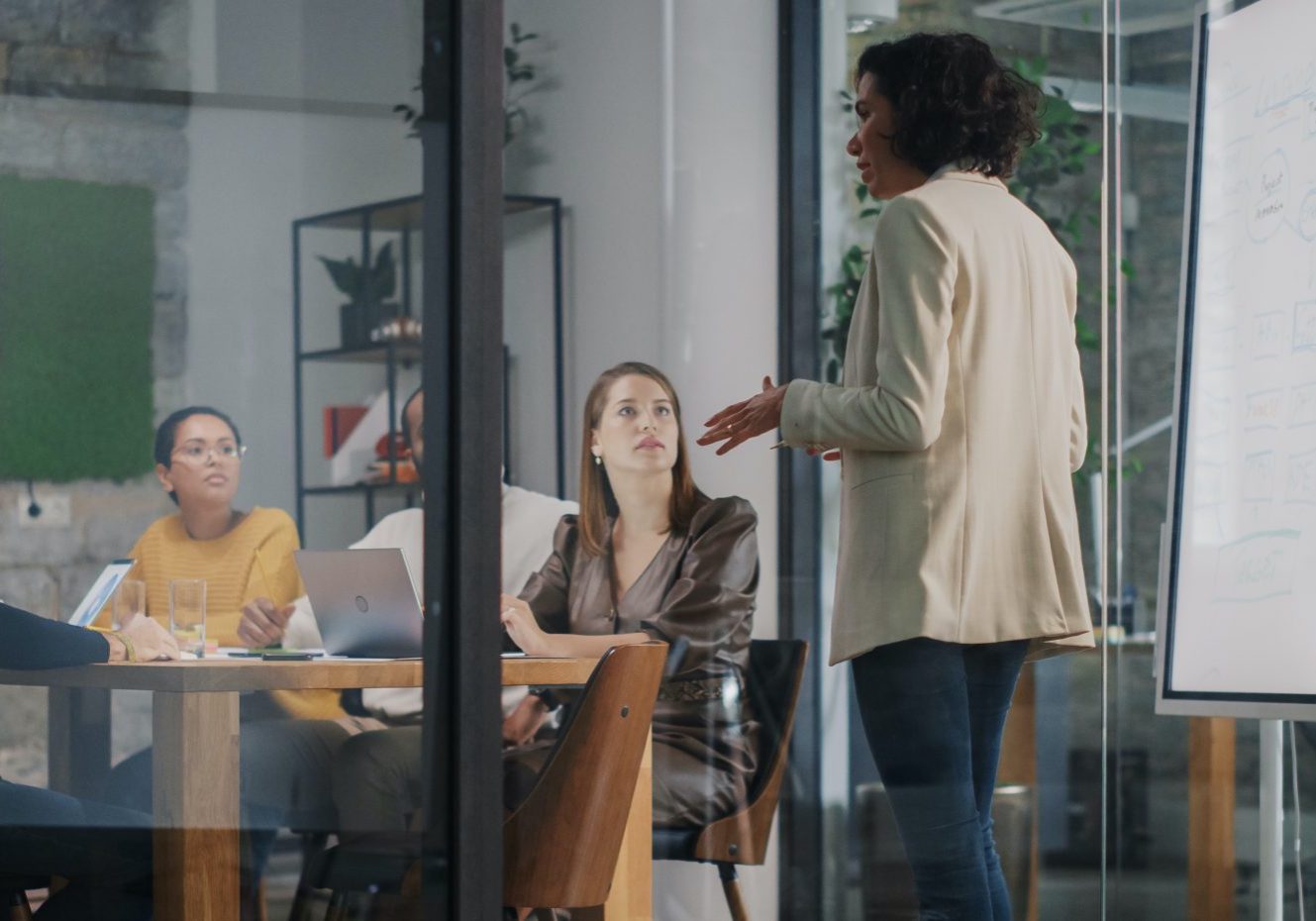 Project Manager Makes a Presentation for a Young Diverse Creative Team in Meeting Room in an Agency. Colleagues Sit Behind Conference Table and Discuss Business Development, User Interface and Design. Project Manager Makes a Presentation for a Young Diverse Creative Team in Meeting Room in an Agency. Colleagues Sit Behind Conference Table and Discuss Business Development, User Interface and Design.
